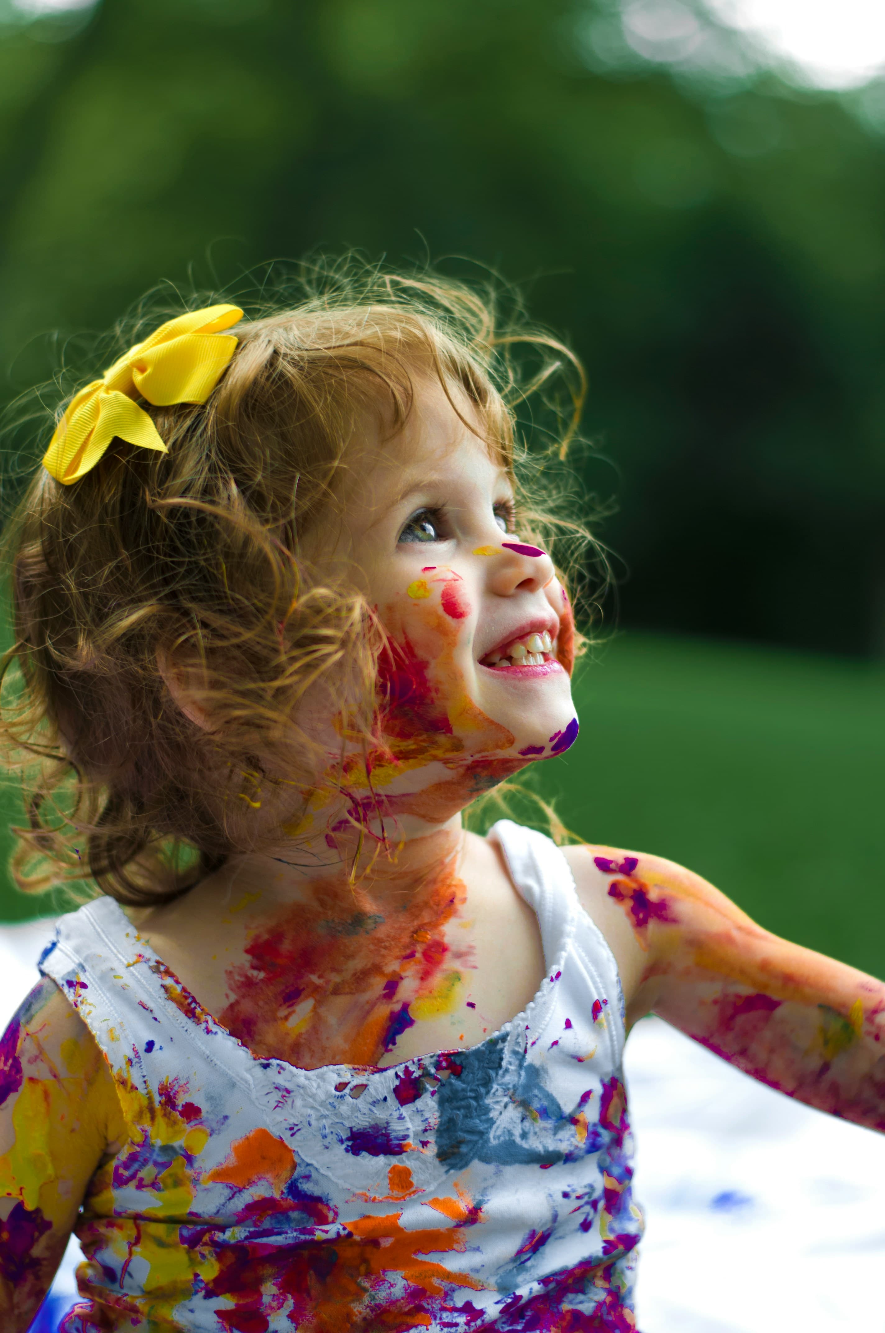 Une petite fille, le visage et les bras couverts de peinture, lève les yeux.