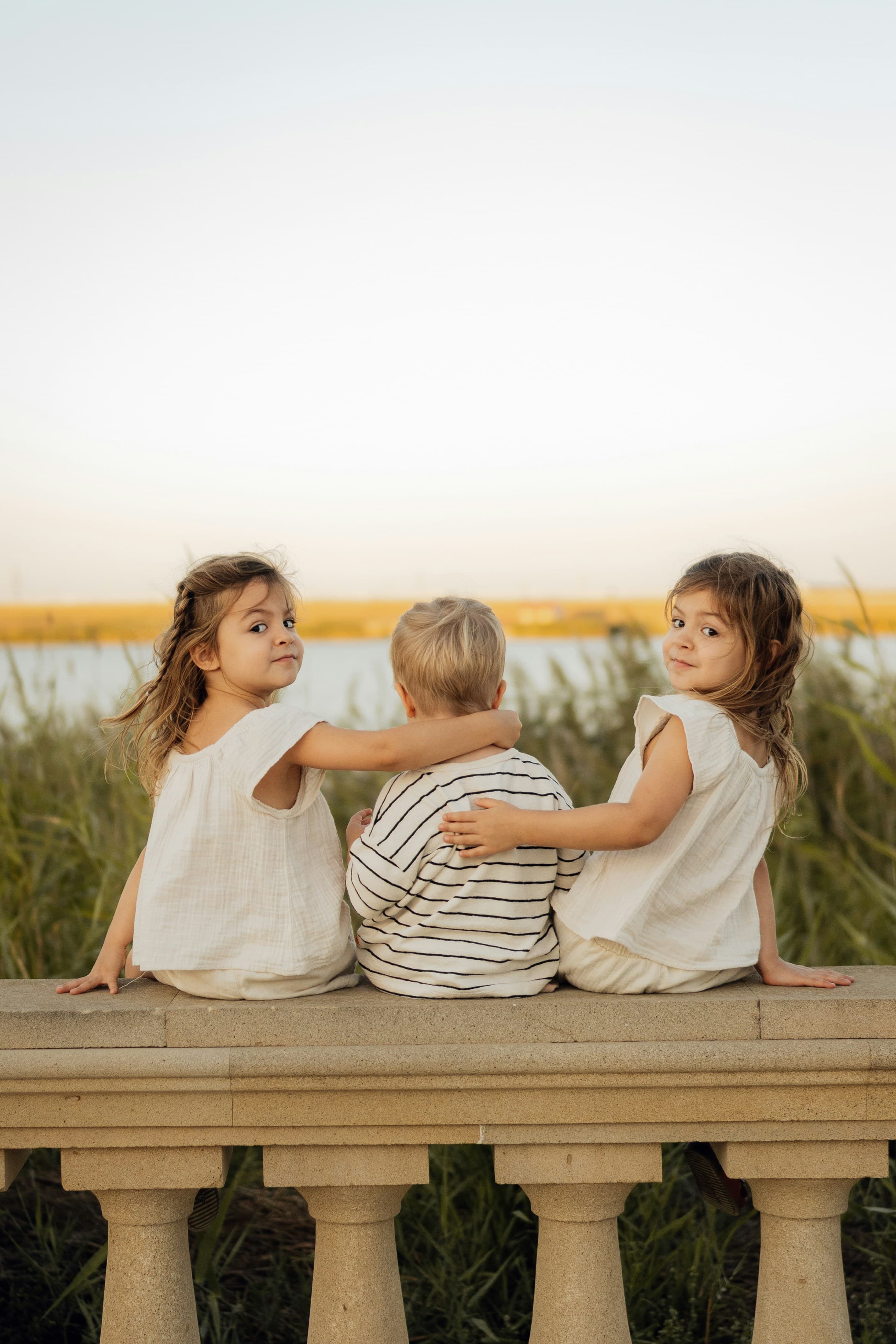 Trois enfants sont assis ensemble sur une balustrade de pierre, avec de l'eau derrière eux.